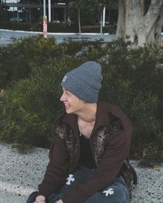 a young man sitting on the ground next to a tree and wearing a beanie