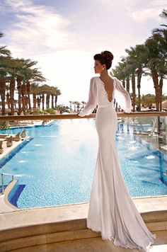 a woman standing in front of a pool wearing a long sleeved wedding dress with open back