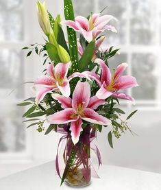 a vase filled with pink lilies and greenery on a table next to a window