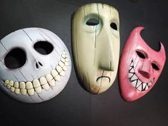 three masks are hanging on the wall with their faces painted white and pink, one has teeth