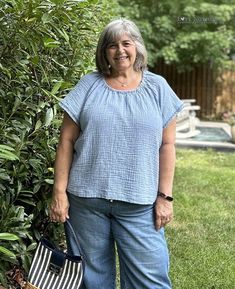 an older woman standing in front of a bush holding a purse and smiling at the camera