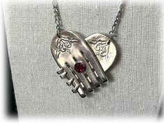 a heart shaped necklace with two hands and a red stone in the middle on a silver chain