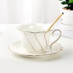 a white tea cup and saucer with a gold stripe on the rim, next to a potted plant