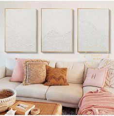 three paintings hang on the wall above a white couch in a living room with pink and gold pillows