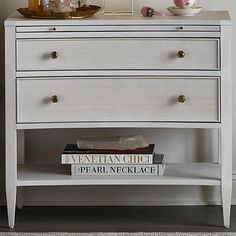 a white dresser with two books on top of it next to a vase and lamp