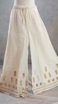 PRODUCT DETAILS This is a beautiful mul-mul cotton Off-White Palazzo Pants with Gota patti borders and gold leather flowers on the Shararaa.. It is beautifully made in soft mul-mul cotton fabric and has a A-Line Flary hem. It also has an attached lining inside. It has a stretchy elastic band and a tying string for a good fit. The Skirt can fit up to size 40 inch waist and can be tightened with the strings for narrower waist. Size & Fit One Size Waist: 28 (expandable to 42) Height: 39.5 inches In Cotton Festival Bottoms, Traditional White Trousers, Elegant Cotton Embroidered Skirt, Traditional White Bottoms For Festive Occasions, Elegant Cotton Skirt With Embroidery, Elegant Embroidered Cotton Skirt, Full Length Cotton Summer Skirt, Fitted Cotton Bottoms For Festivals, Full Length Cotton Skirt For Summer