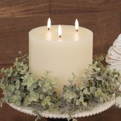 a white candle sitting on top of a cake plate with greenery around the edge