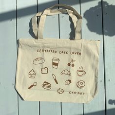 Measuring 20 inches by 15 inches, our Certified Cafe Lover screen-printed tote bag is made from 100% cotton canvas, ensuring durability and style. It comes with an outer zipper and a convenient mini pocket inside with another zipper for added functionality. Adorned with charming cafe items such as utensils, cake, pastries, and coffee, this bag is perfect for carrying your essentials while showcasing your passion for all things cafe. Carry it with pride and let your love for cafes shine with our Coffee Clothes Aesthetic, Cafe Tote Bag, Coffee Shop Merch, Cafe Items, Screen Print Tote Bag For Shopping, Cute Tote Bag Design, Tote Bag Design Ideas, Artsy Shopping Tote Bag, Kawaii Tote Bag With Cute Design