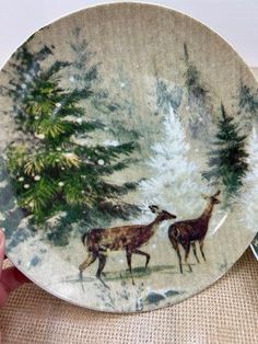 a hand holding a plate with two deers in the woods on it and snowing trees