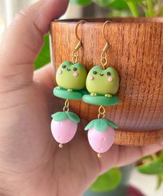 a pair of green and pink frog earrings on top of a wooden planter next to a potted plant