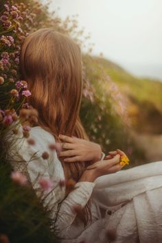 a woman sitting in the grass with her back to the camera and looking at flowers