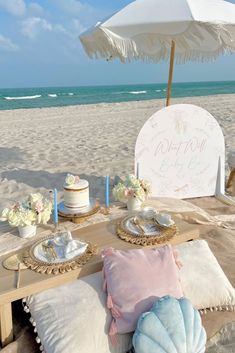 a table set up for a baby shower on the beach with an umbrella over it