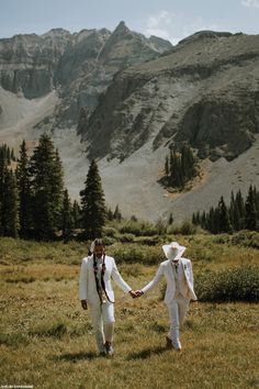 two people in white suits holding hands and walking through the grass with mountains in the background