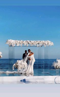 a bride and groom standing in front of an arch with white flowers on the beach