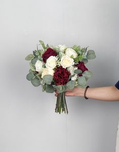 a person holding a bouquet of flowers in their hand on a gray background with white, red and green foliage