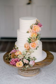 a three tiered white cake with pink and orange flowers on the top is sitting on a wooden platter