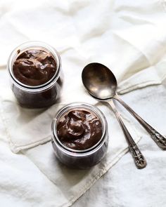 two jars filled with chocolate pudding on top of a white cloth next to spoons