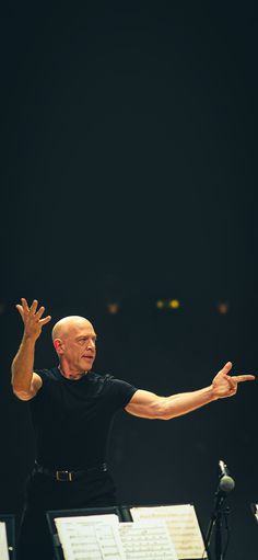 an older man with his arms outstretched in front of music sheets and microphones on stage