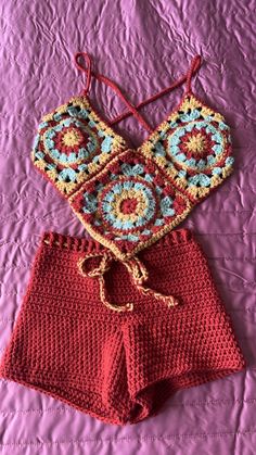 two pieces of crocheted clothing are laying on a pink bed spread, one has an open top and the other has short shorts