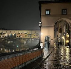 people are walking down an alley way at night with lights on and water running through the cobblestone