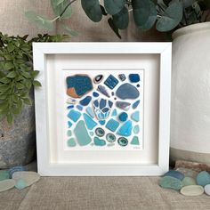 a white frame with blue and green sea glass in it next to a potted plant