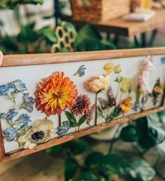 a person holding up a wooden frame with flowers on it and plants in the background