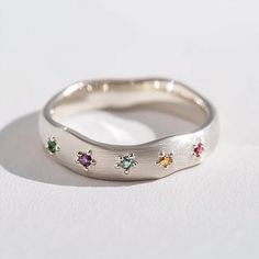 a silver ring with five different colored stones on it's side, sitting on a white surface