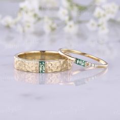 two gold wedding rings with green stones on them and white flowers in the back ground