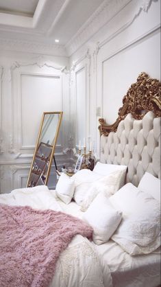 a white bed topped with lots of pillows next to a gold framed mirror on the wall