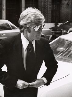 a man in a suit and tie standing next to a car with his hands on his hips