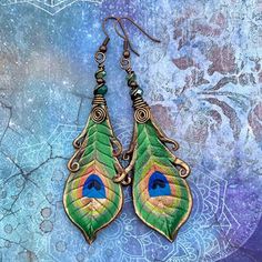 two pairs of peacock feathers are hanging from earrings on a blue and purple patterned background