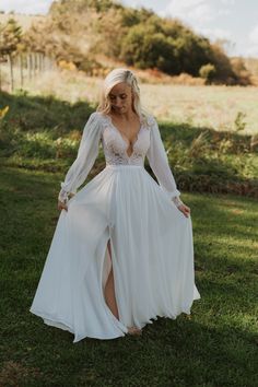 a woman standing in the grass wearing a white dress with long sleeves and high slit