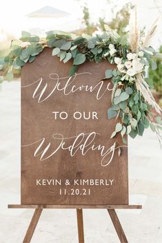 a welcome sign with greenery and flowers is displayed on a easel for an outdoor ceremony