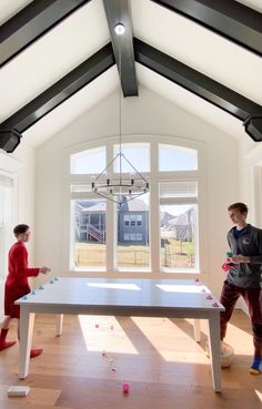 two boys are playing ping pong in an open room with large windows and wood flooring