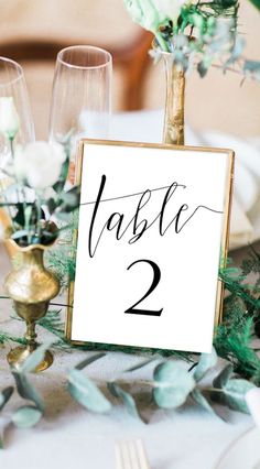 the table numbers are displayed in gold vases with greenery on them, along with wine glasses and silverware