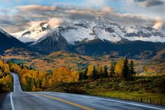 an empty road in the mountains with snow capped mountains on either side and trees lining both sides