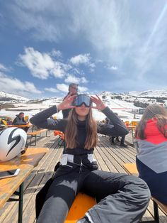 a woman sitting on top of a wooden bench wearing ski goggles and holding her hands up to her face