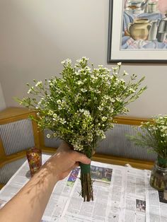 a person holding flowers on top of a table next to a vase with white flowers