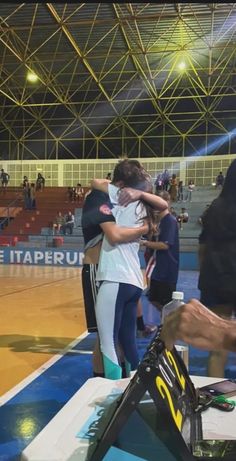 two people hug each other on the sidelines of an indoor basketball court with fans in the background