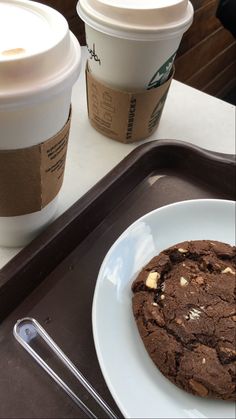 two cups of coffee sit next to a chocolate cookie on a tray with a plate