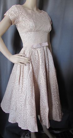 "Pretty lace party dress in a buttercream tone of beige.  Typical of late'50s, early '60s design, it has a tightly fitted empire bodice and full skirt. The waist is set slightly lower than the natural waist and is highlighted with a self fabric belt with 6\" bow at front. It has 4 1/2\" cap sleeves. The back plunges 5 1/2\" to 15\" metal zipper. The under bust seam is 9 1/2\" from throat. The dress is lined. No maker label Size: no size label--35\" bust; 27\" at the natural waist; 34\" at slight Beige A-line Lace Dress, Beige Feminine Vintage Dress For Party, Beige Vintage Dress For Party, Cream A-line Lace Dress, Beige Party Dress With Scalloped Lace, Beige Scalloped Lace Party Dress, Fitted Beige A-line Lace Dress, Beige Fitted A-line Lace Dress, Feminine Cream Vintage Party Dress