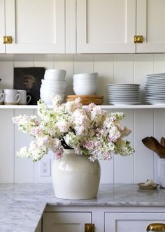 a white vase filled with lots of flowers on top of a counter next to plates