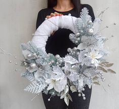 a woman is holding a wreath with silver and white flowers on it, while standing against a wall