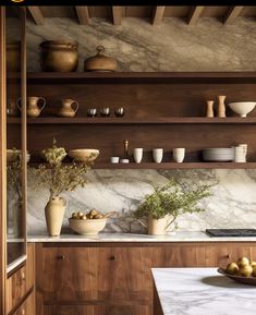 a kitchen with marble counter tops and wooden shelves filled with bowls, cups, vases and fruit