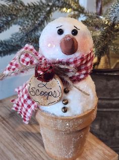 a snowman sitting in an ice cream cone on top of a wooden table next to a christmas tree