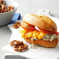 a chicken sandwich with tomatoes, cheese and onion on a table next to pretzels