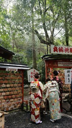 two women in kimonos standing next to each other near a brick wall and trees