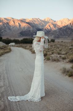 a woman in a white dress and hat standing on a dirt road with mountains in the background