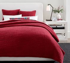 a bed with red bedspread and pillows in a white walled room next to a night stand