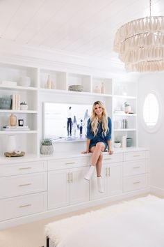 a woman sitting on top of a kitchen counter next to white cupboards and shelves